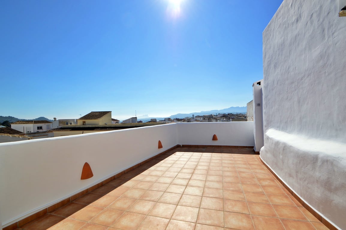 Terraza en la azotea con suelo de baldosas de terracota y muros blancos, ofreciendo vistas despejadas al pueblo y las montañas bajo el sol mediterráneo.