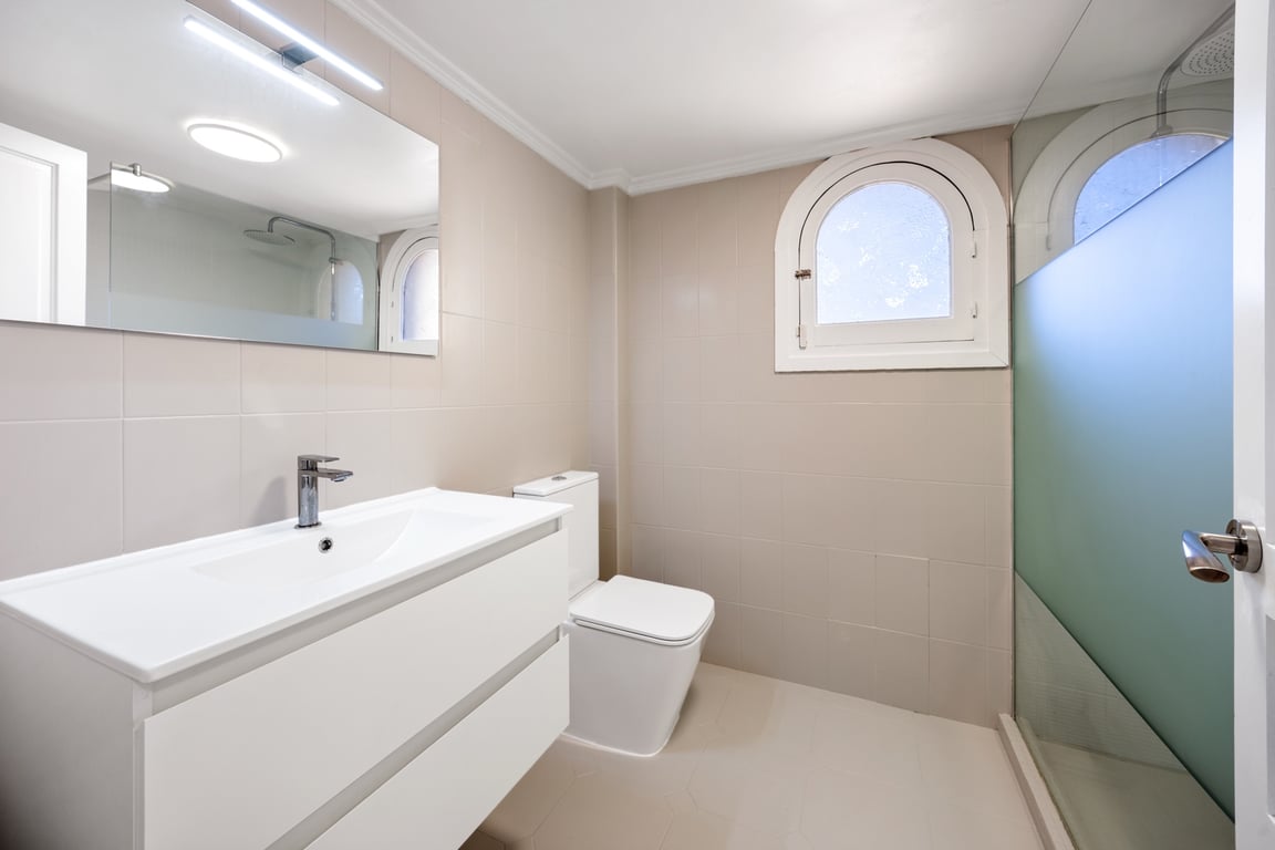Baño moderno con lavabo suspendido blanco, ducha con mampara de cristal y una ventana de arco característica. Paredes alicatadas en tonos neutros y luz LED sobre el espejo.