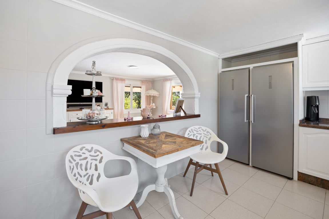 Cocina con arco abierto hacia el salón, equipada con frigorífico doble de acero inoxidable y zona de desayuno con mesa de madera y sillas de diseño.