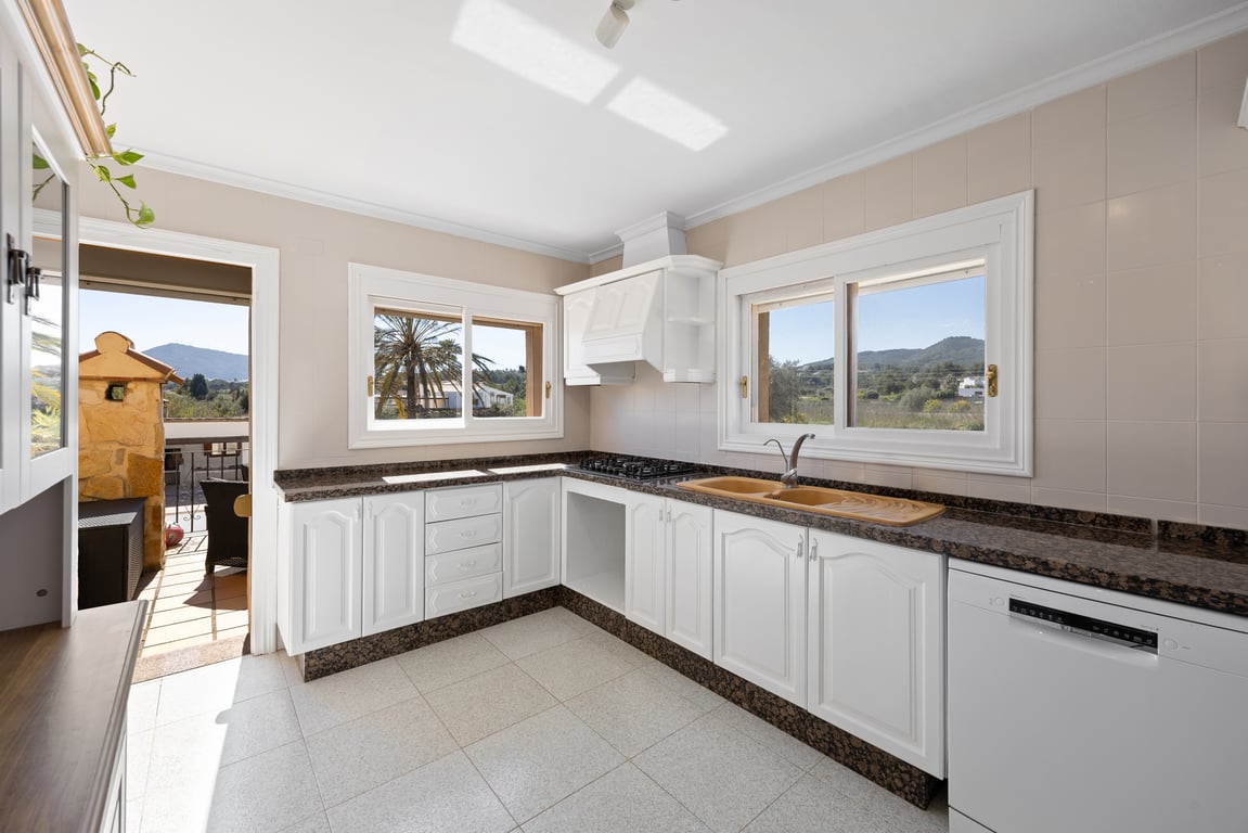 Cocina luminosa con armarios blancos, encimera de granito oscuro y vistas despejadas al campo. Incluye acceso directo a la terraza y equipamiento funcional.