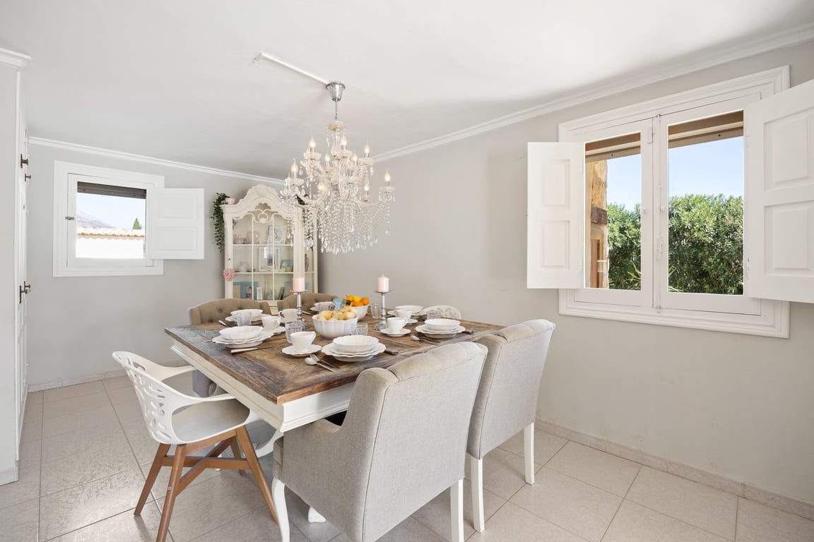 Comedor luminoso con mesa de madera rústica, sillas tapizadas y una elegante lámpara de araña. Las ventanas con contraventanas blancas ofrecen luz natural y vistas al jardín.