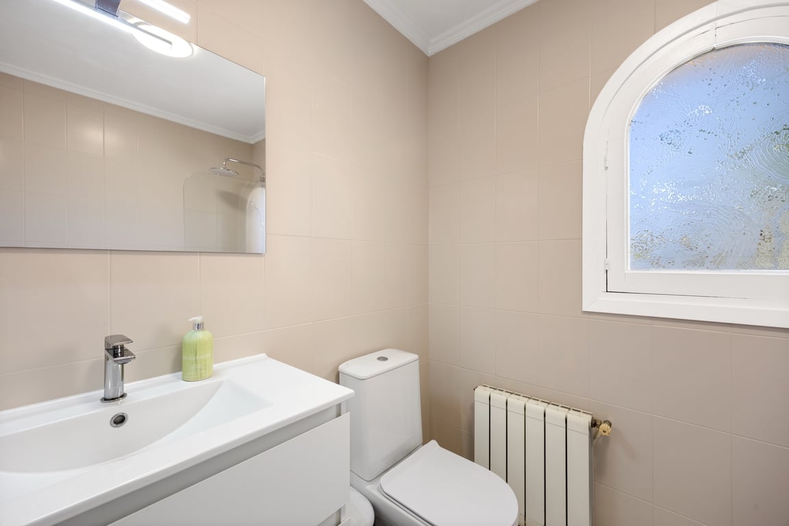 Cuarto de baño funcional con azulejos neutros, lavabo con mueble blanco, radiador y una ventana de arco con cristal translúcido que aporta luz natural.