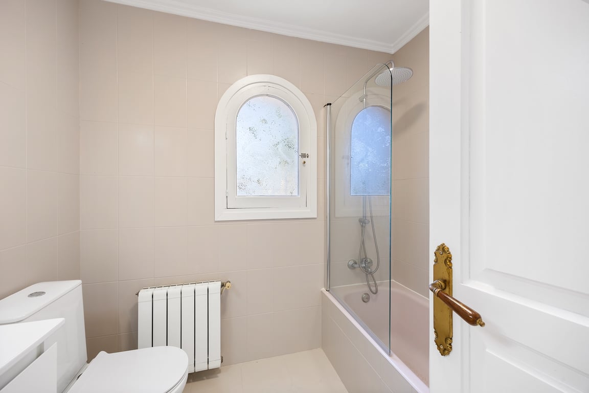 Cuarto de baño funcional con bañera, mampara de cristal y una característica ventana de arco. Incluye calefacción por radiador y acabados en tonos neutros.