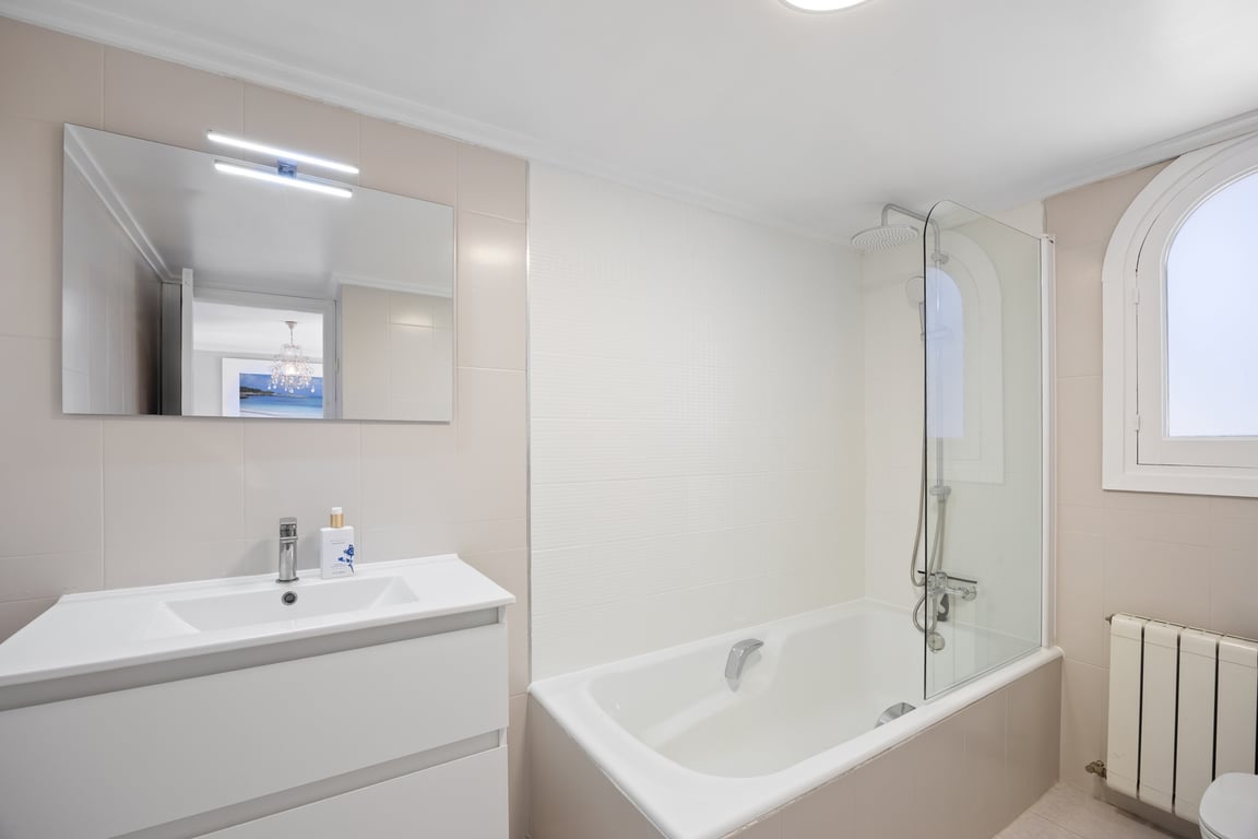 Cuarto de baño moderno con lavabo blanco, espejo iluminado y bañera con mampara de cristal. Incluye una ventana de arco que aporta luz natural y azulejos en tonos neutros.