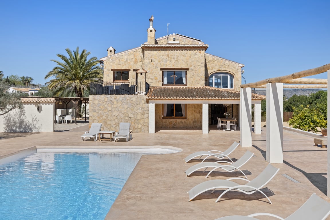 Fachada de piedra natural con naya cubierta, amplia terraza de gres y piscina. La villa muestra su arquitectura de dos niveles y zonas de descanso exteriores bajo el sol.