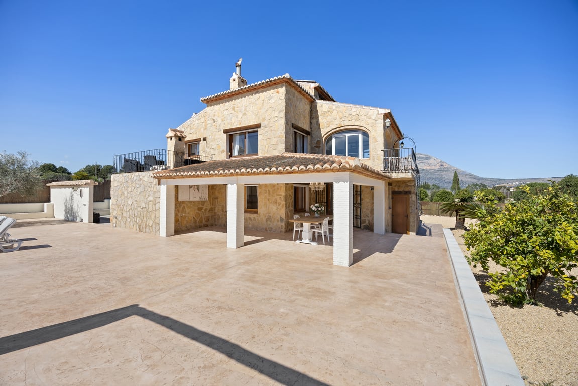 Fachada de piedra natural con naya cubierta de pilares blancos, amplia terraza pavimentada y vistas despejadas al Montgó bajo un cielo despejado en Jávea.