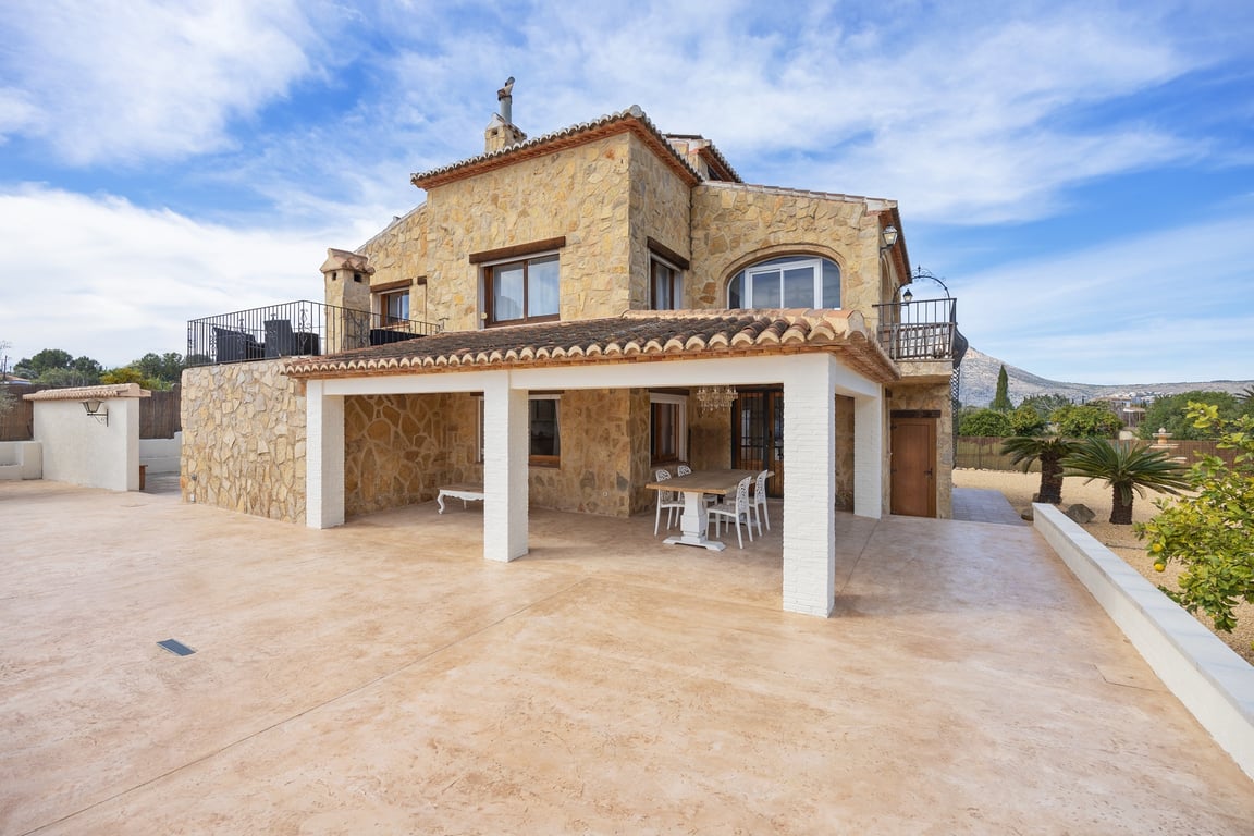 Fachada de piedra natural con naya cubierta, pilares blancos y terrazas superiores, integrada en un amplio espacio exterior pavimentado con entorno mediterráneo.