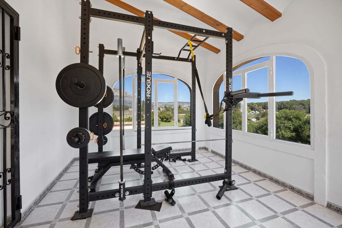Gimnasio privado equipado con rack profesional Rogue, ubicado en una estancia luminosa con vigas de madera y vistas despejadas al entorno natural de Jávea.