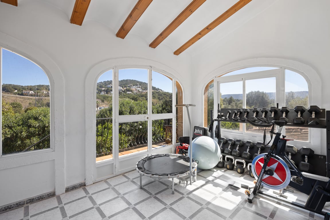 Naya acristalada utilizada como gimnasio, con vigas de madera, suelo cerámico y vistas despejadas a la montaña a través de sus arcos tradicionales.