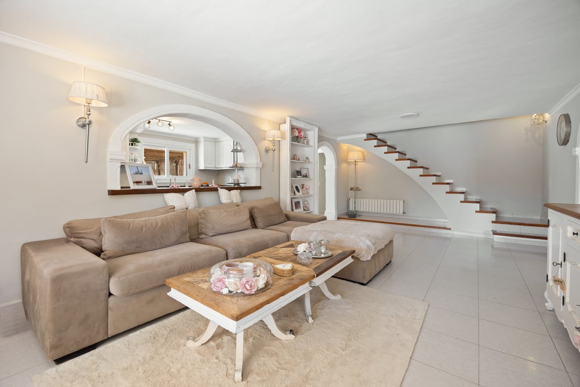 Salón espacioso con suelos cerámicos, sofá rinconero y un arco decorativo que conecta con la cocina. Destaca la escalera de diseño con peldaños de madera que sube a la planta superior.