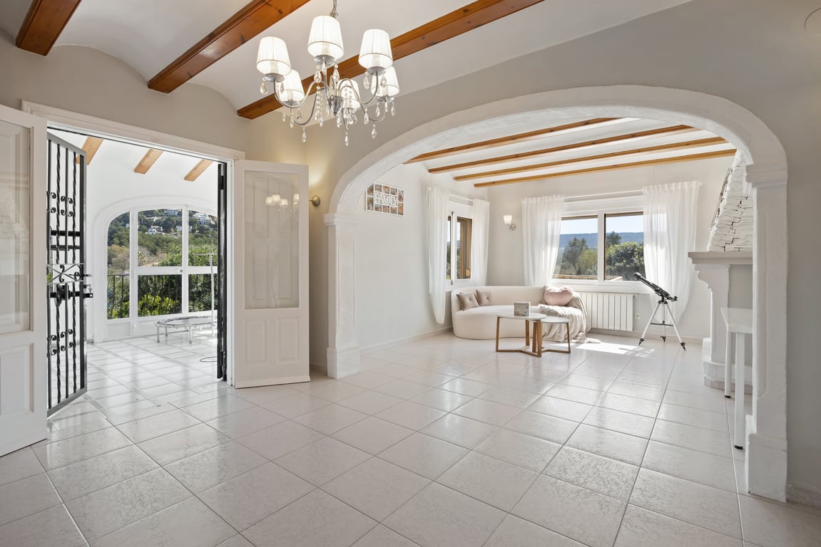 Salón luminoso con vigas de madera, chimenea de piedra blanca y un gran arco que conecta con una zona de estar con sofá curvo y vistas al exterior.