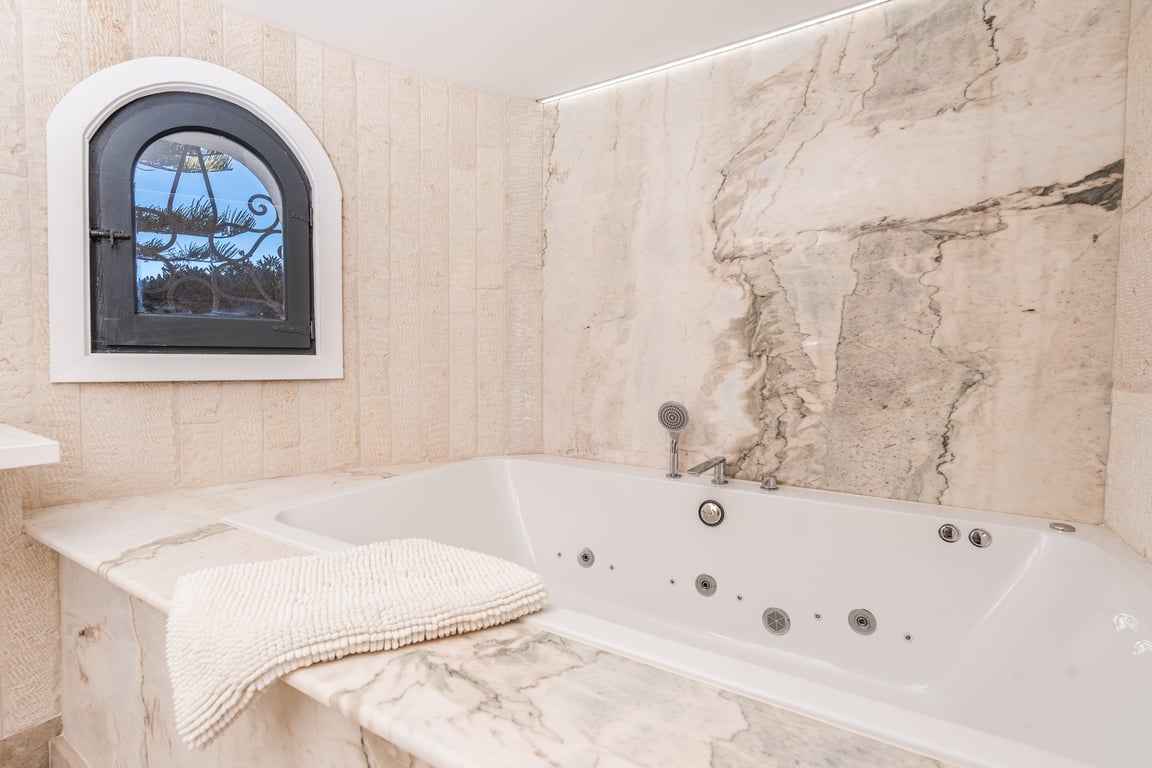 Bañera de hidromasaje integrada con revestimiento de mármol veteado, paredes de piedra natural y una ventana de arco característica.