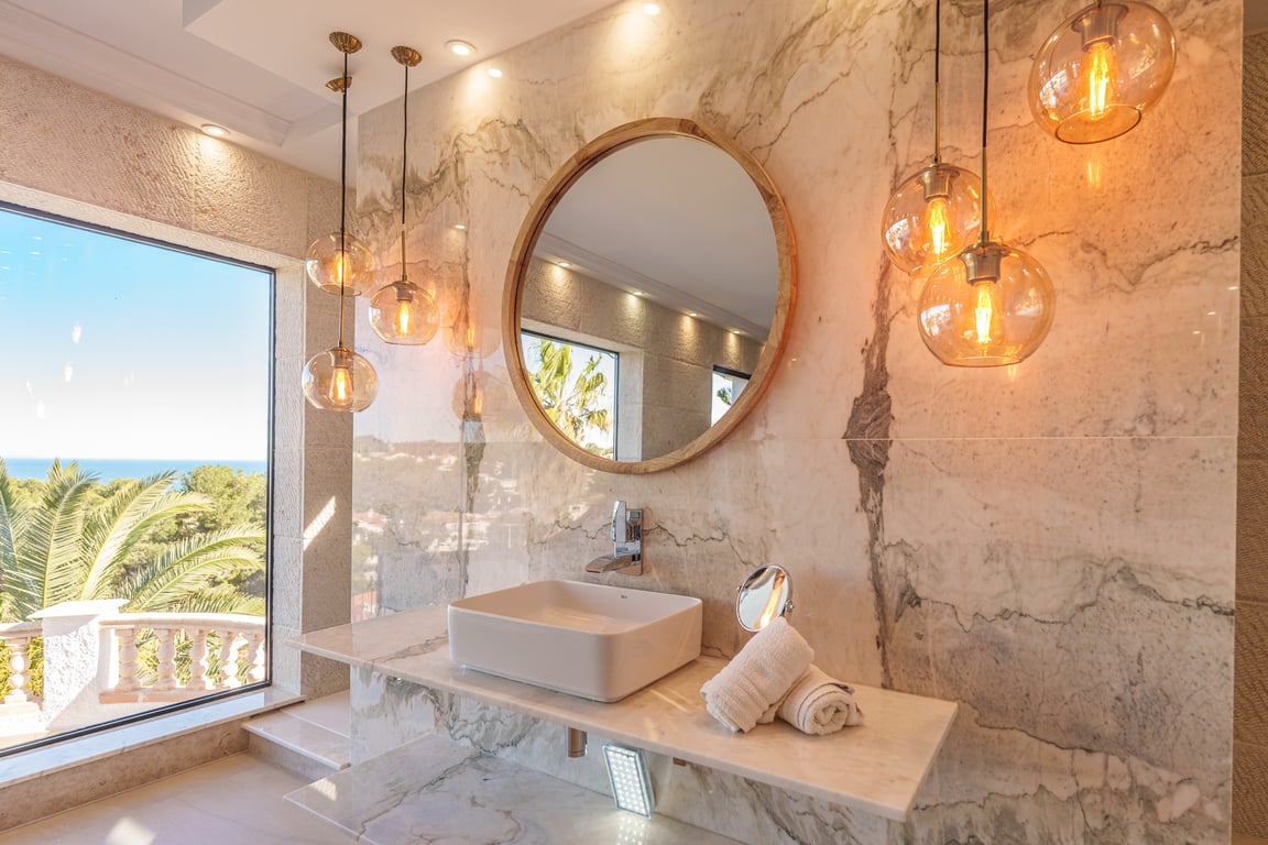 Baño de la suite principal con paredes de mármol veteado, lavabo sobre encimera, espejo circular de madera y lámparas colgantes de cristal con vistas al mar.