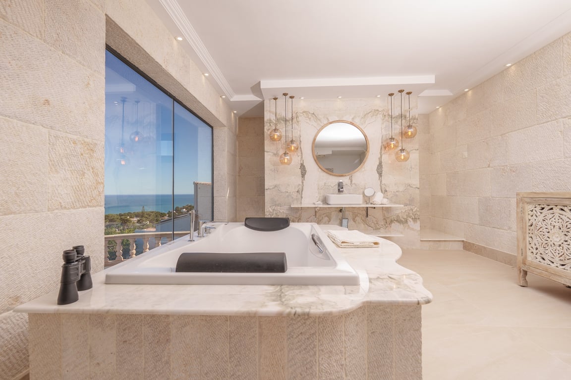 Baño de la suite principal con paredes de piedra caliza, bañera encastrada en mármol con vistas al mar y lavabo de diseño con iluminación colgante decorativa.