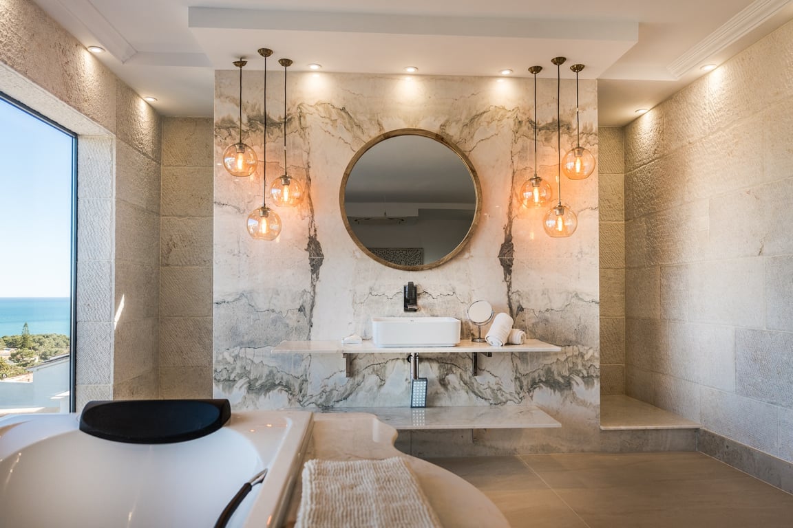 Baño de la suite principal con paredes de piedra natural, encimera de mármol veteado, lámparas colgantes esféricas y vistas despejadas al mar.