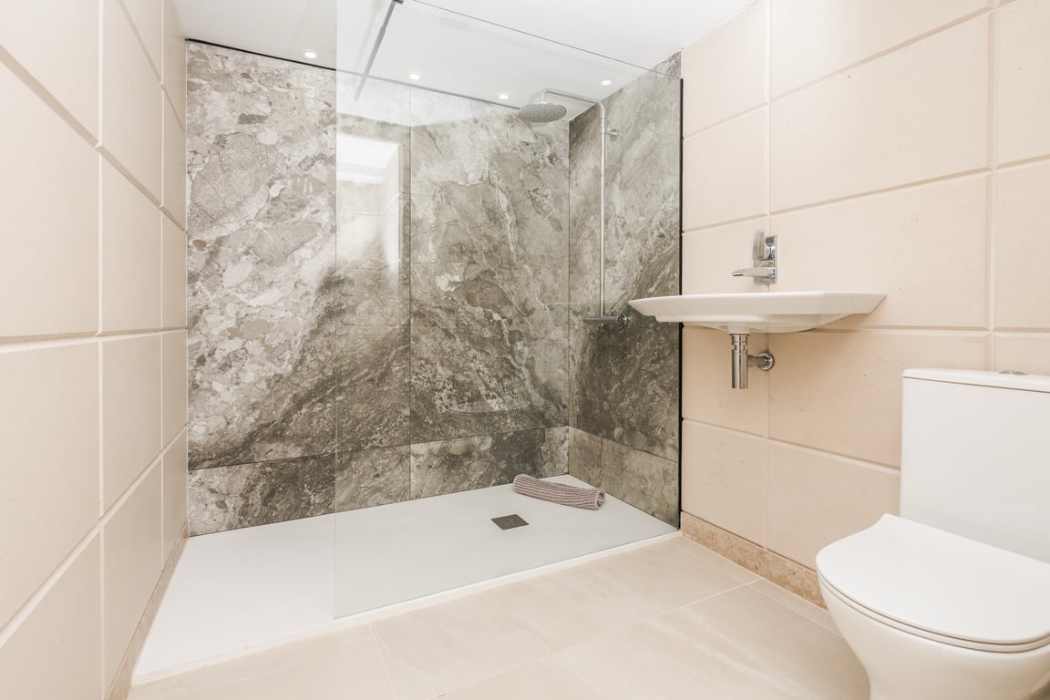 Baño moderno con ducha a ras de suelo, mampara de cristal y pared de mármol gris. Incluye sanitarios blancos minimalistas y azulejos en tonos crema.
