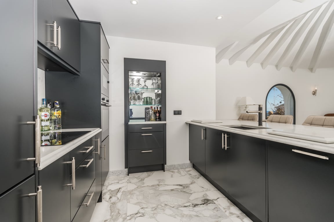 Cocina de diseño con mobiliario oscuro, encimeras de mármol y suelos coordinados. Destaca la isla central y el mueble bar empotrado bajo un techo con vigas blancas radiales.