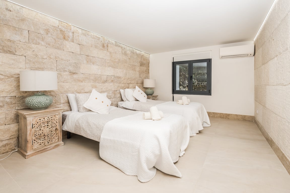 Dormitorio con dos camas individuales, pared de piedra natural texturizada, mesitas de madera tallada e iluminación LED integrada en el techo.
