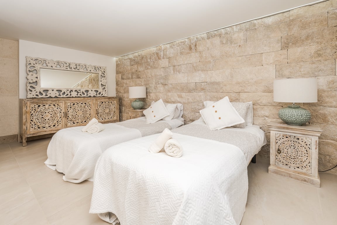 Dormitorio con dos camas individuales, pared de piedra natural texturizada y mobiliario de madera tallada artesanalmente. Incluye iluminación integrada y suelos de tono neutro.
