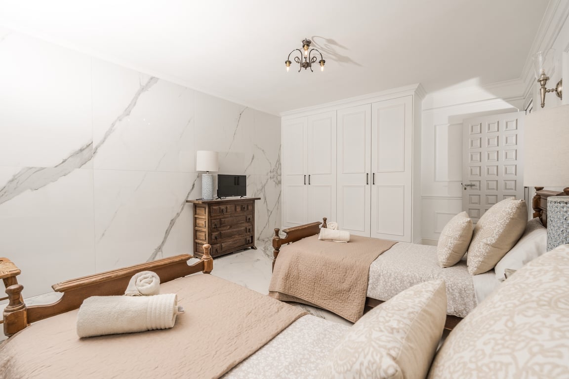 Dormitorio con dos camas individuales, pared revestida de mármol veteado, armarios empotrados blancos y cómoda de madera rústica con televisión.