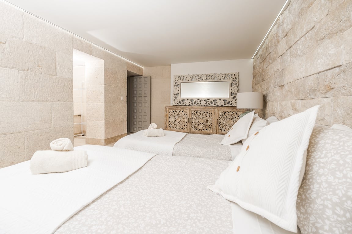 Dormitorio con paredes de bloques de piedra caliza, dos camas individuales y muebles de madera tallada. Dispone de iluminación LED indirecta integrada en el techo.