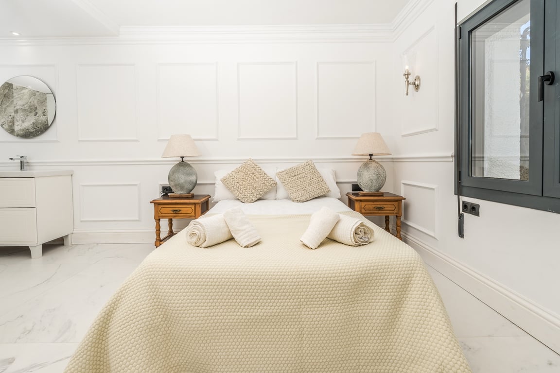 Dormitorio con paredes de molduras blancas y suelo de mármol. Incluye un área de tocador integrada con espejo circular y mesitas de noche de madera clásica.
