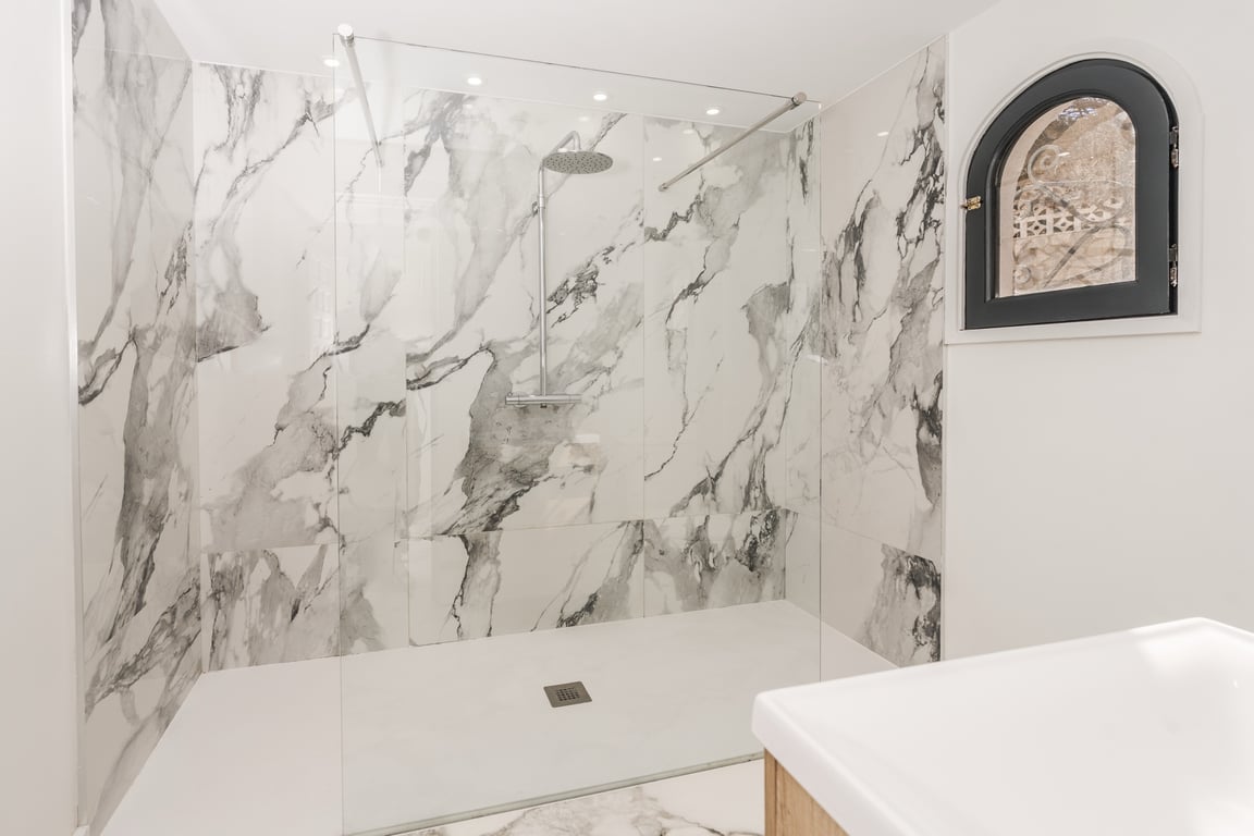 Ducha de efecto mármol con mampara de cristal fija, grifería cromada y una ventana de arco con marco negro que aporta luz natural y carácter mediterráneo.