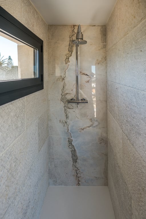 Ducha de obra con pared central de mármol veteado y laterales de piedra caliza texturizada. Incluye grifería cromada y una ventana que aporta luz natural al espacio.