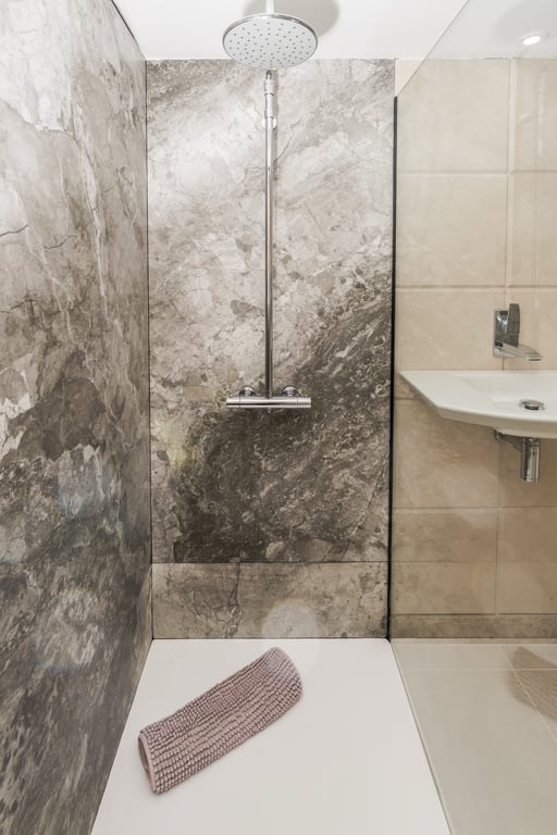 Ducha walk-in con revestimiento de mármol gris, cabezal de lluvia cromado y plato de ducha blanco, integrada en un baño de diseño contemporáneo.