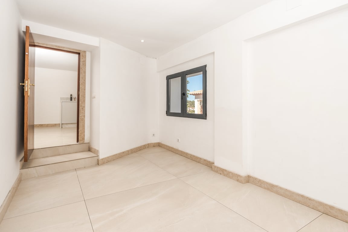 Habitación luminosa con suelos de piedra caliza, zócalos a juego y paredes blancas. Incluye una ventana de marco oscuro y un acceso elevado hacia una estancia contigua.