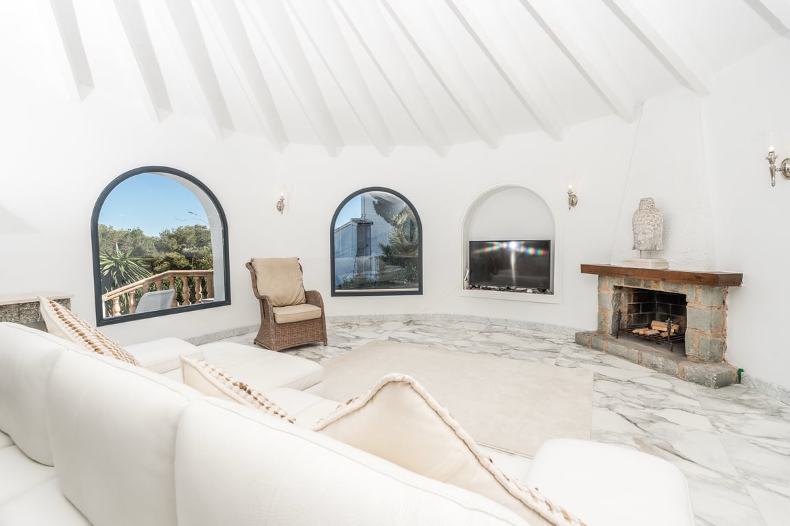 Salón circular con techo de vigas radiales blancas, suelo de mármol y chimenea de piedra. Los ventanales de arco ofrecen vistas al exterior en un ambiente luminoso.