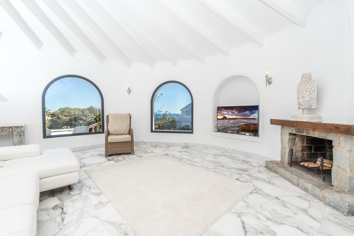 Salón circular con techos de vigas blancas, suelo de mármol y chimenea de piedra rústica. Las ventanas en arco ofrecen vistas despejadas al entorno natural y al mar.
