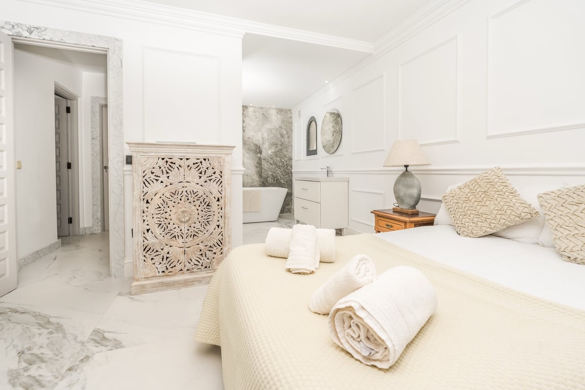 Suite principal con baño abierto, suelos de mármol y una pared de piedra natural tras la bañera exenta. Destaca el mobiliario tallado y las elegantes molduras de pared.