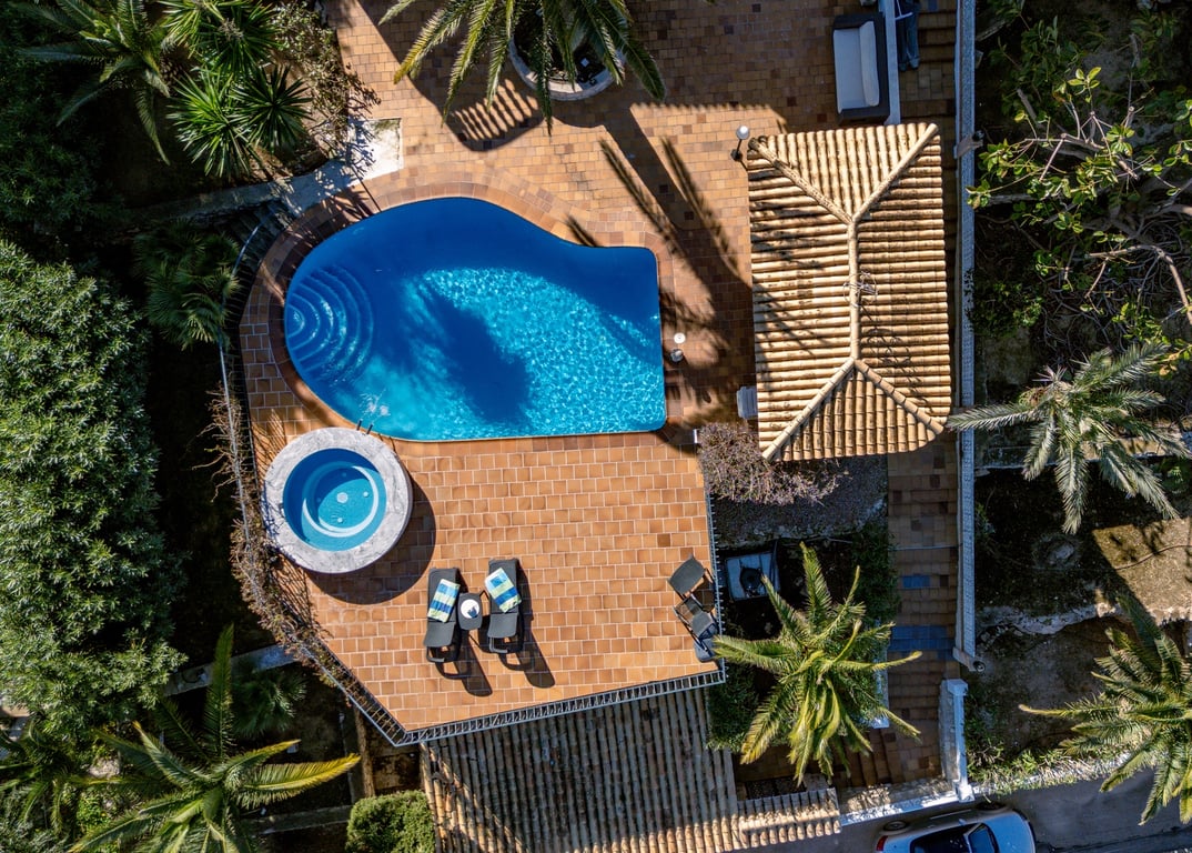 Vista aérea de la zona exterior con piscina de forma orgánica, jacuzzi circular y amplia terraza de gres rodeada de palmeras y vegetación mediterránea.