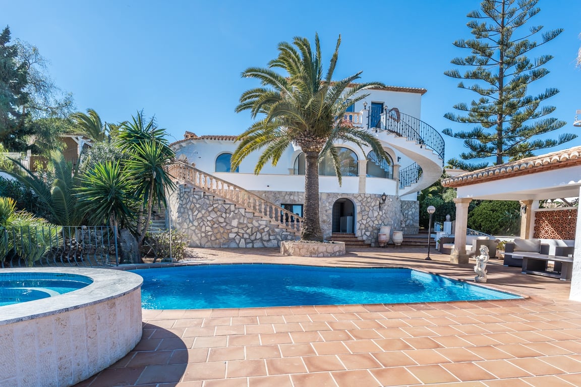 Vista exterior con piscina, jacuzzi circular y fachada de piedra. Destaca la escalera de caracol que conecta las plantas entre palmeras y jardines mediterráneos.