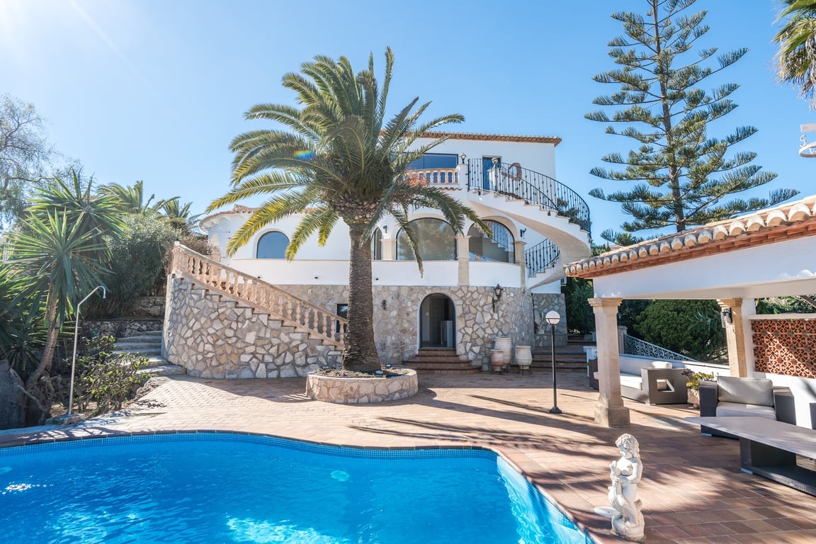 Vista exterior de la villa con piscina, destacando la fachada de piedra natural, las escaleras curvas y el jardín mediterráneo con palmeras bajo el sol de Jávea.