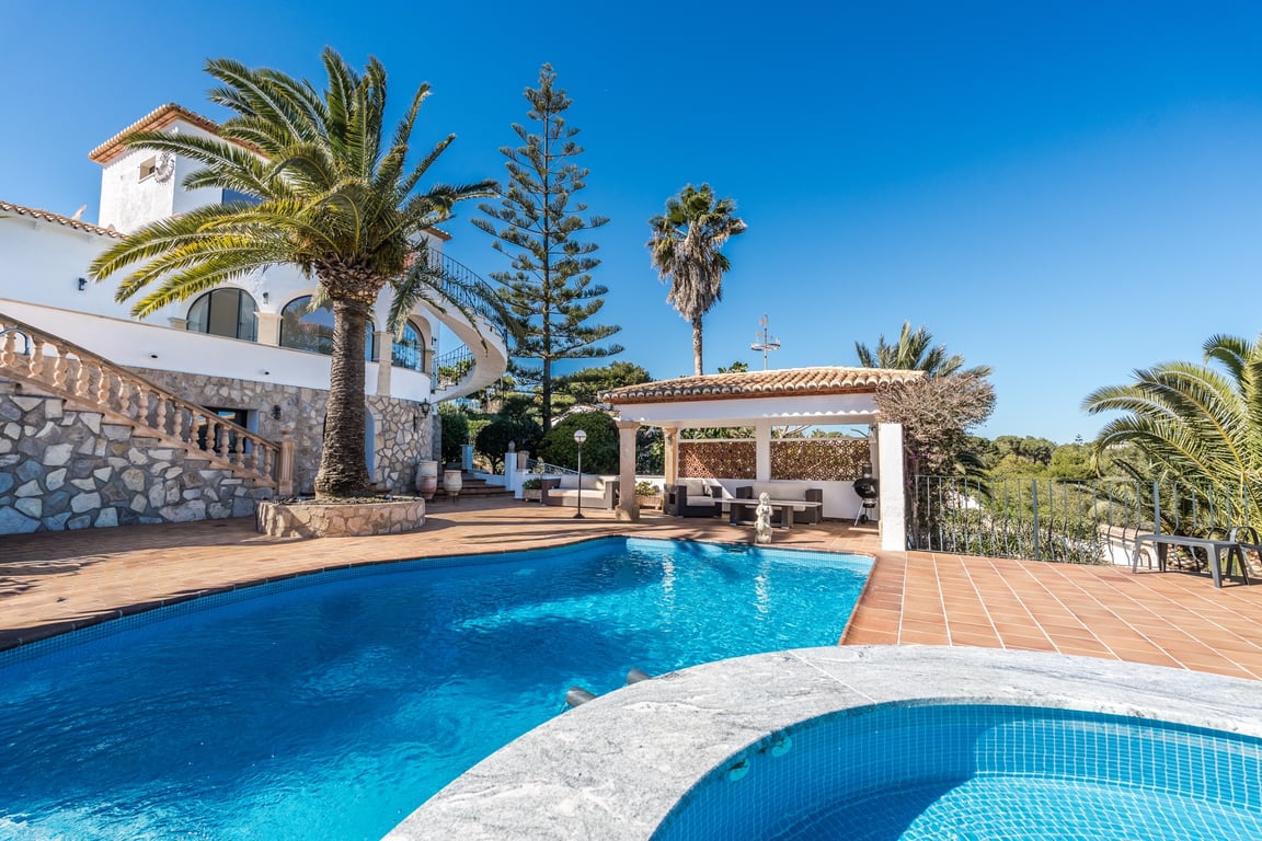 Zona exterior con piscina y jacuzzi, rodeada de una terraza de terracota, palmeras y una fachada de piedra natural con escalera clásica y porche cubierto.