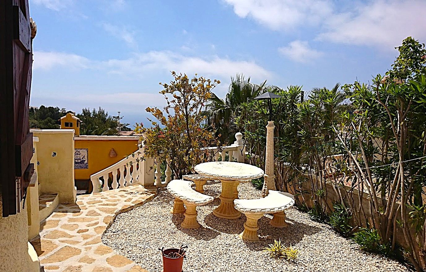 Área de descanso exterior con mesa y bancos de piedra tallada sobre suelo de grava y piedra natural, rodeada de vegetación mediterránea y vistas despejadas.
