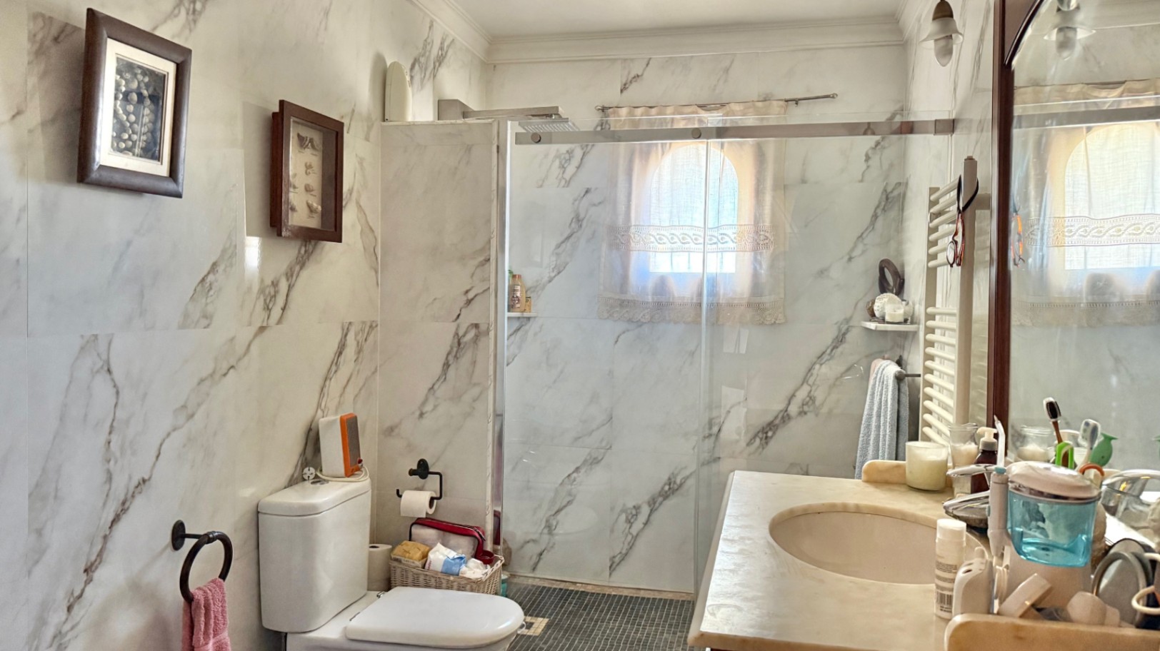 Baño con paredes revestidas en mármol, ducha de obra con mampara de cristal y ventana de arco. Incluye lavabo integrado y radiador toallero.