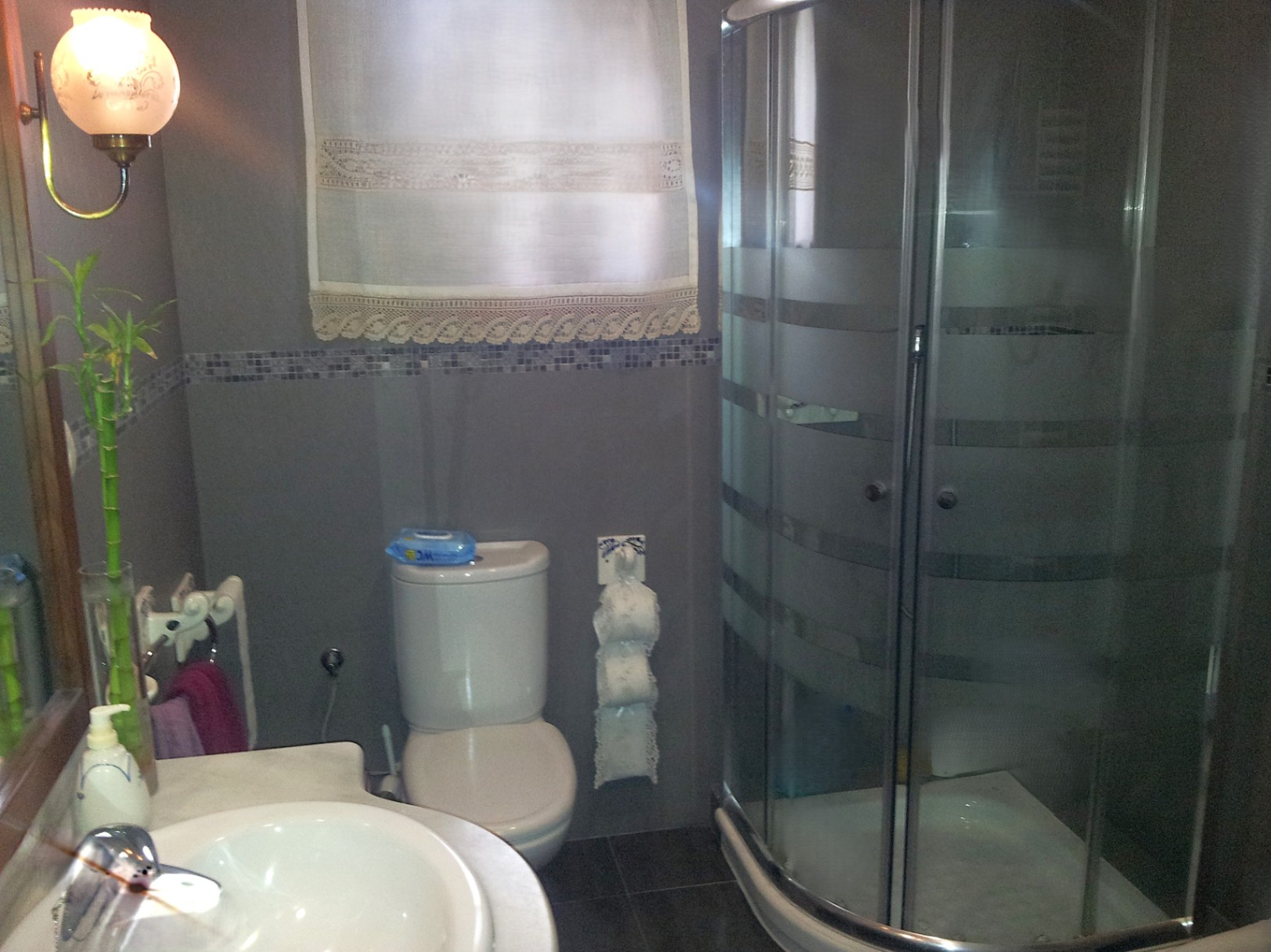 Baño funcional con ducha de esquina con mampara curva, paredes en tonos grises con cenefa de mosaico y encimera de mármol en el lavabo.