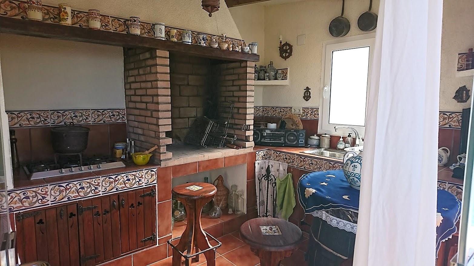 Cocina de verano rústica con barbacoa de obra, encimeras decoradas con azulejos tradicionales, fogones de gas y armarios de madera tallada.