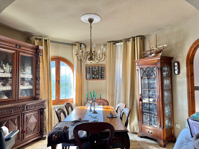 Comedor de estilo rústico con muebles de madera maciza tallada, vitrina para cristalería y mesa central bajo una lámpara de araña de latón y luz natural.
