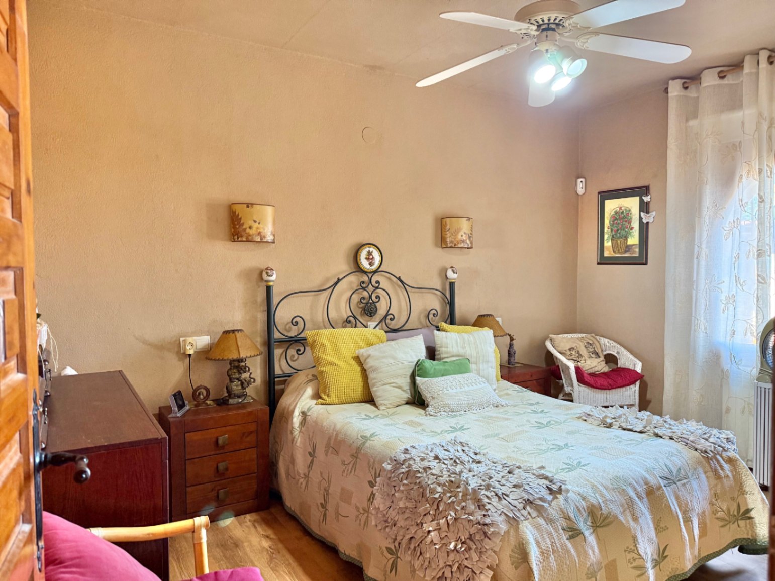 Dormitorio con cabecero de forja, mesitas de madera y ventilador de techo. La estancia cuenta con suelos de madera y luz natural a través de ventanales con cortinas.
