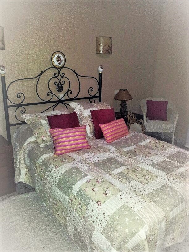 Dormitorio con cama doble, cabecero de hierro forjado negro y colcha de estilo patchwork. Incluye un sillón de mimbre blanco y paredes en tonos neutros.