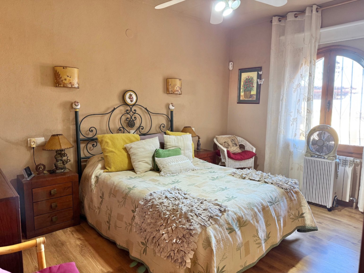 Dormitorio rústico con cabecero de forja, suelos de madera y ventana de arco. Dispone de ventilador de techo, radiador y mobiliario de madera clásica.