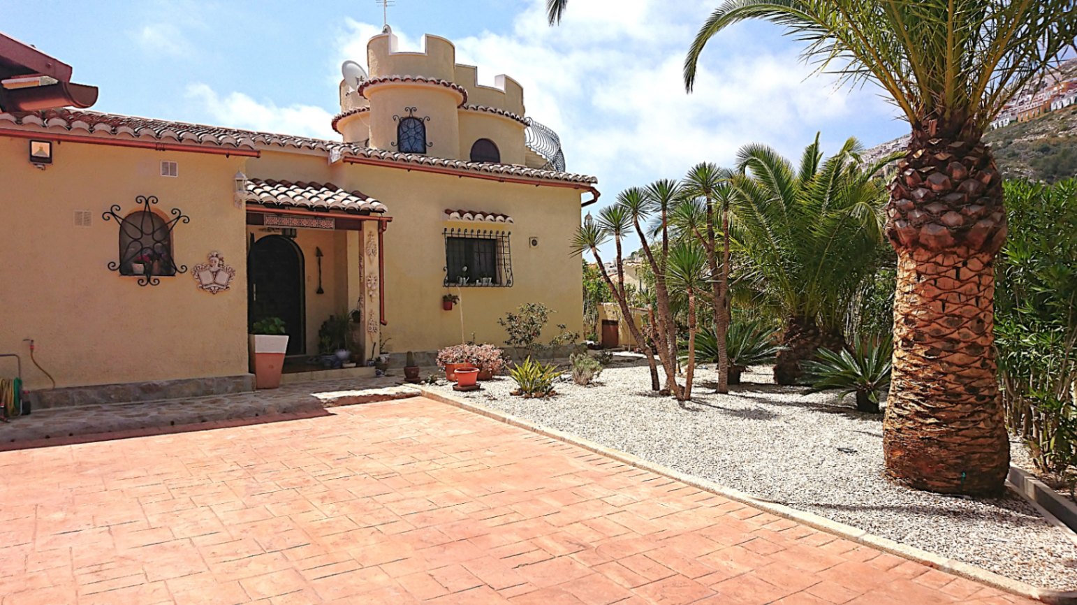 Fachada exterior con torre almenada, entrada principal bajo porche de tejas y jardín mediterráneo con palmeras y grava.