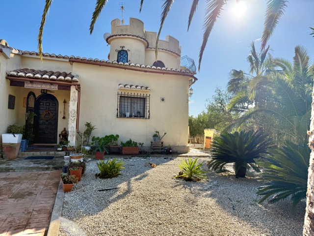 Fachada exterior con torre almenada, entrada principal con puerta de madera tallada y jardín de grava con vegetación mediterránea bajo luz natural.