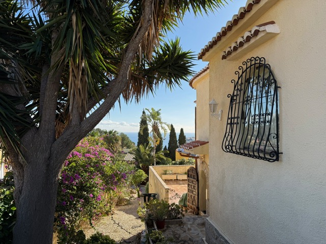 Fachada exterior con ventana de rejería artesanal, rodeada de vegetación mediterránea y buganvillas, con vistas despejadas hacia el mar.