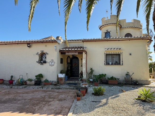Fachada exterior de la villa con torreón almenado, ventanas de arco con rejas de hierro forjado y entrada principal con puerta de madera y porche de teja.