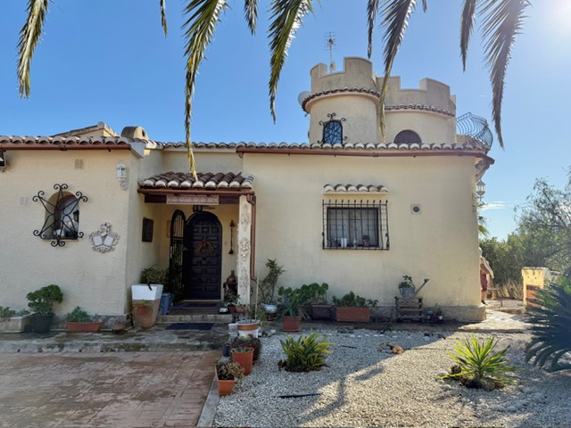 Fachada principal con torre característica, entrada de madera tallada y rejería artesanal en un entorno de jardín mediterráneo con plantas crasas.